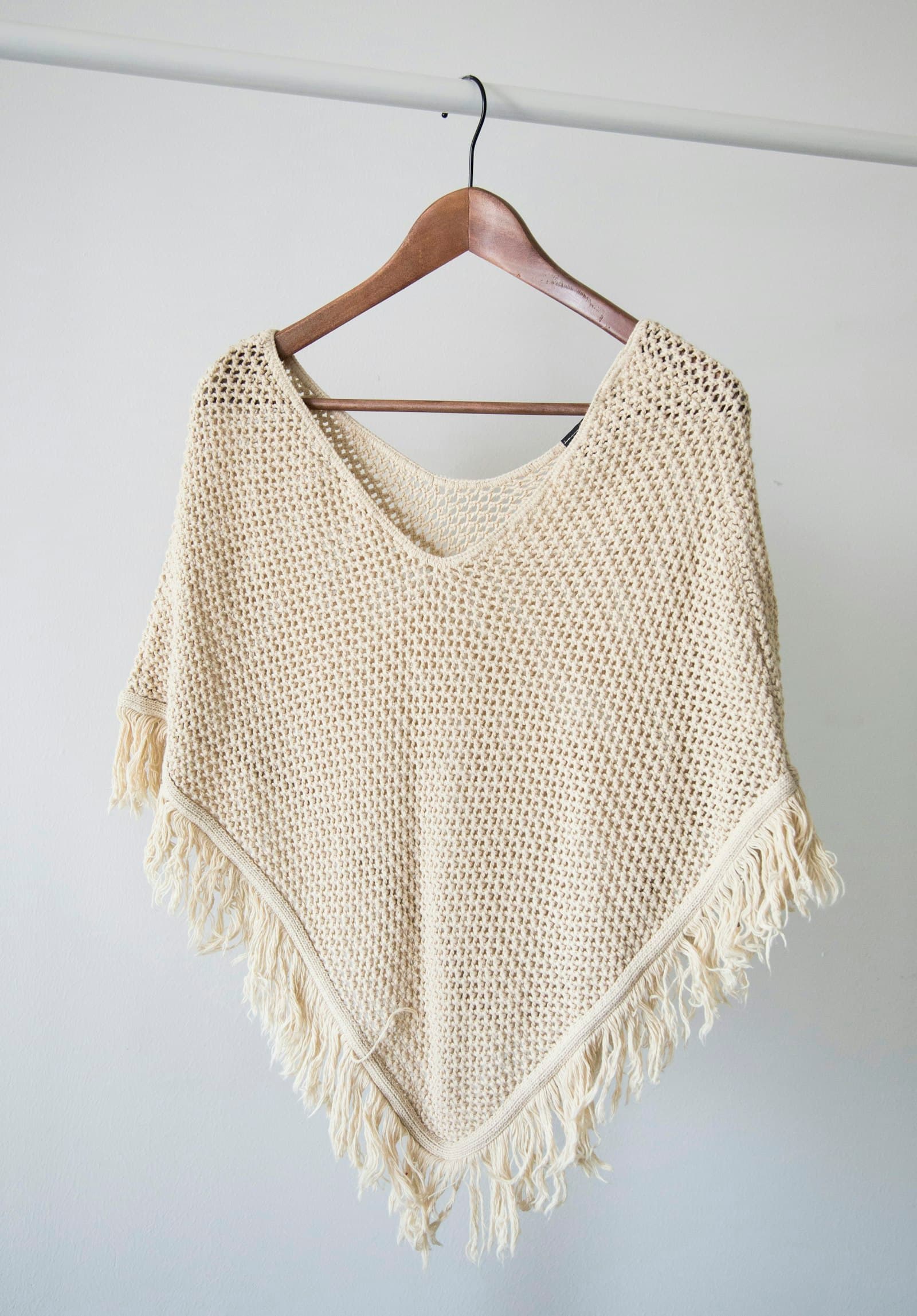 Cream crochet poncho with fringe hanging on a wooden hanger